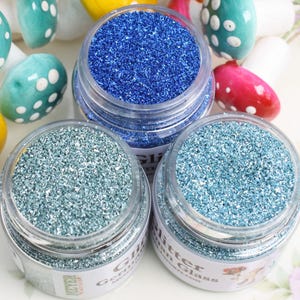 Pode incluir: Três pequenos frascos de vidro cheios de glitter azul, azul-verde e azul claro. Os frascos têm etiquetas brancas com o texto "Glitter in Glass" impresso neles.