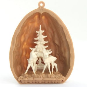 Op de afbeelding: Een klein, gesneden houten ornament in de vorm van een walnootschelp. In de schelp bevindt zich een miniatuur scène met twee witte herten die onder een kleine, witte dennenboom staan.