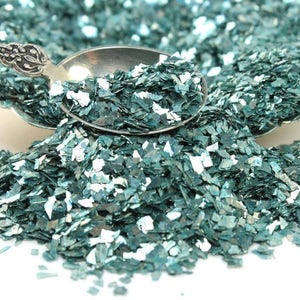 May include: Teal iridescent glitter flakes in a silver spoon. The glitter is scattered on a white surface.
