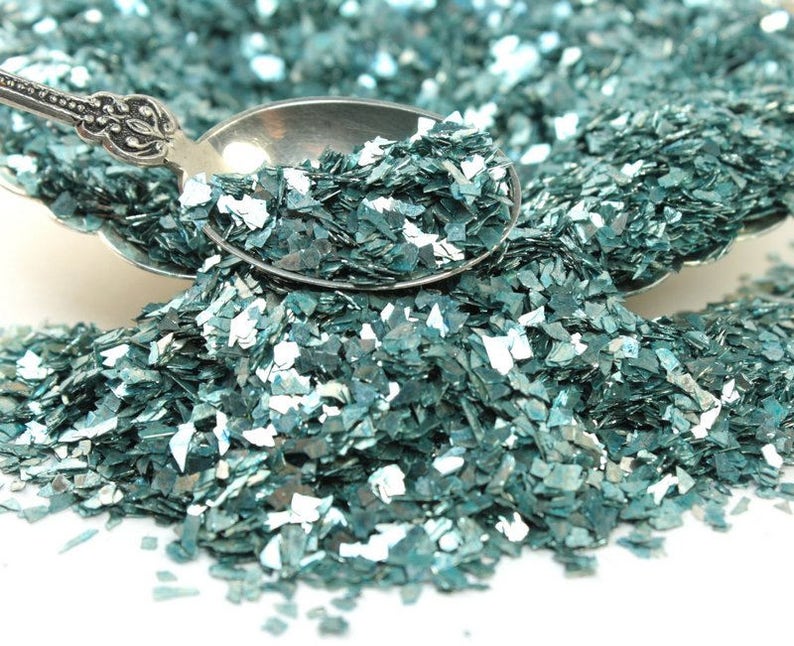 May include: Teal iridescent glitter flakes in a silver spoon. The glitter is scattered on a white surface.