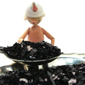 May include: A wooden figurine wearing a white hat pours black glitter into a silver spoon. The glitter is also scattered on a silver tray.