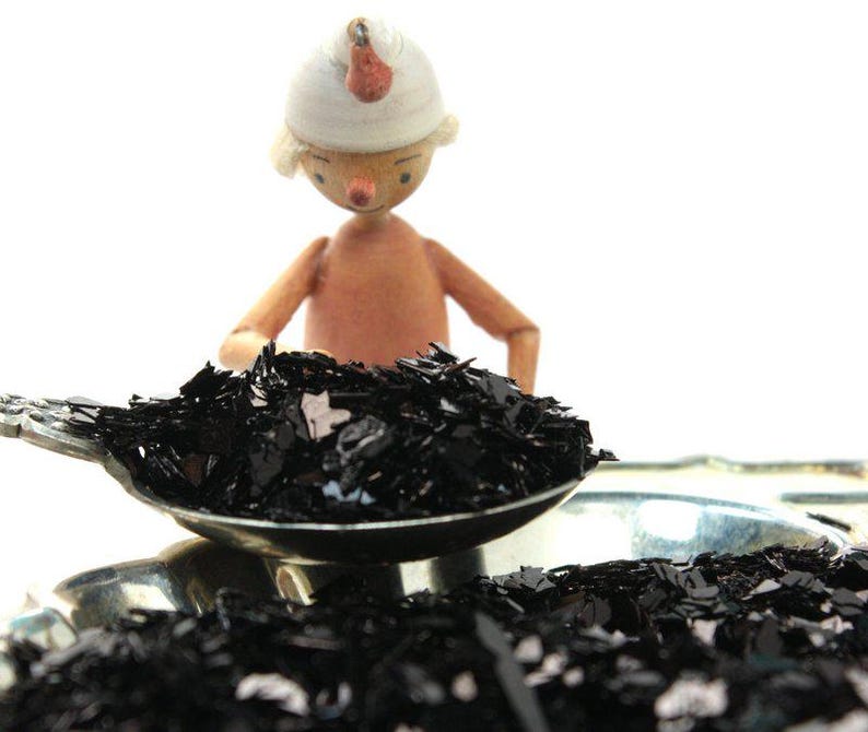 May include: A wooden figurine wearing a white hat pours black glitter into a silver spoon. The glitter is also scattered on a silver tray.