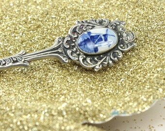 May include: A silver spoon with an ornate handle and a blue and white porcelain inset featuring a windmill design. The spoon is resting on a bed of gold glitter.