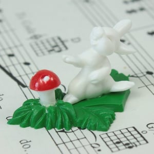 May include: A white rabbit figurine with a red mushroom on a green leaf base. The figurine is standing on a sheet of music.