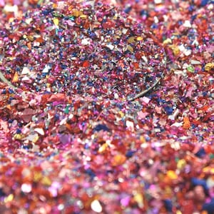 May include: A close-up of a pile of iridescent glitter in shades of pink, red, blue, and silver. The glitter is scattered on a surface and is illuminated by a light source.