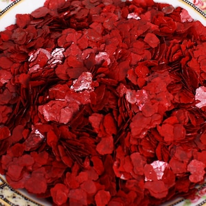 May include: A close-up of a bowl filled with red mica flakes. The flakes are small and irregular in shape, and they have a shiny, metallic finish.