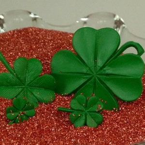 May include: Four green four-leaf clovers of varying sizes on a red glitter background.