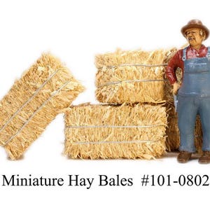 May include: A set of four miniature hay bales, made of straw, with a miniature farmer figurine standing in front of them. The farmer is wearing a red shirt, blue overalls, and a brown hat. The hay bales are tied with twine. Miniature Hay Bales #101-0802