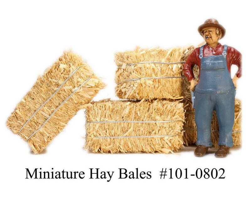 May include: A set of four miniature hay bales, made of straw, with a miniature farmer figurine standing in front of them. The farmer is wearing a red shirt, blue overalls, and a brown hat. The hay bales are tied with twine. Miniature Hay Bales #101-0802
