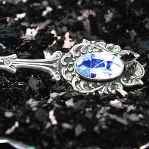 May include: A silver spoon with an ornate design and a blue and white porcelain inlay. The spoon is resting on a bed of black glitter.