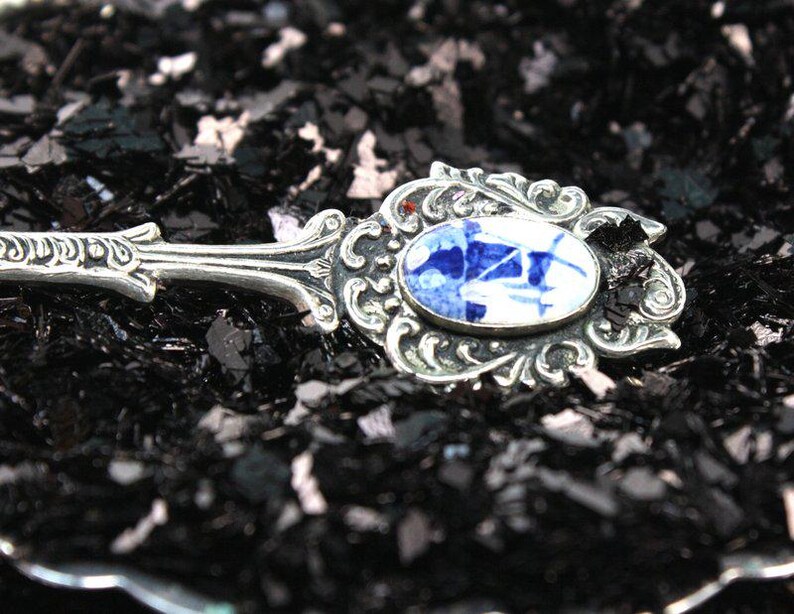 May include: A silver spoon with an ornate design and a blue and white porcelain inlay. The spoon is resting on a bed of black glitter.