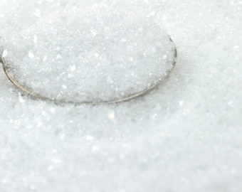 May include: A close-up of a spoon filled with white iridescent glitter. The glitter is scattered around the spoon, creating a sparkling effect.