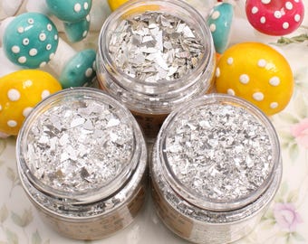 May include: Three small glass jars filled with silver glitter flakes. The jars are on a white surface with colorful mushroom decorations.