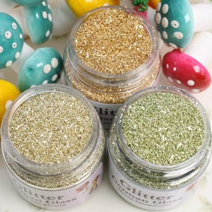 Pode incluir: Três pequenos frascos de vidro cheios de glitter dourado, prateado e verde. Os frascos têm etiquetas que dizem "Glitter German Glass".