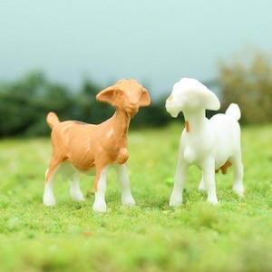 May include: Two miniature goat figurines. One is tan with white legs, the other is white. They stand on a bed of green grass, with a blurred green and blue background.