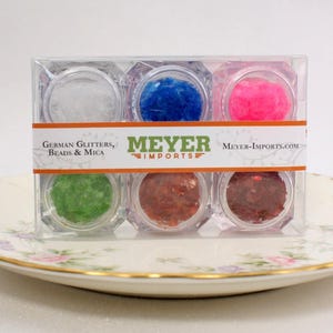 Może przedstawiać: Zestaw sześciu małych słoiczków wypełnionych kolorowym brokatem, oznaczonych "German Glitters, Beads & Mica" i "Meyer Imports".