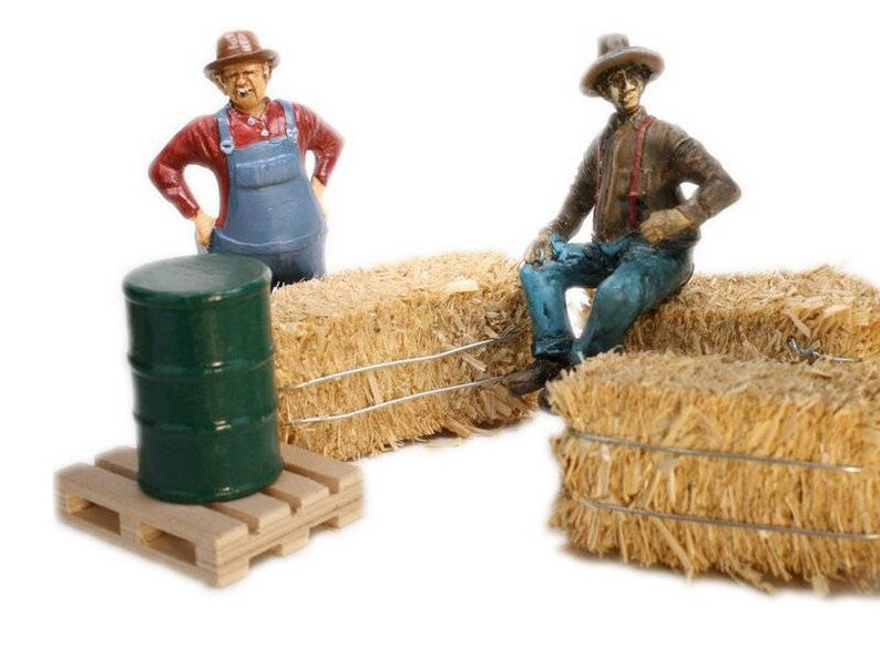 May include: Two miniature figurines of farmers, one wearing a straw hat and overalls, the other wearing a straw hat and blue jeans, sitting on bales of hay. A green barrel sits on a wooden pallet.