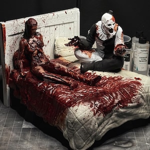 Puede incluir: Un diorama detallado representa una escena espantosa en una cama, con una figura cubierta de sangre y un personaje parecido a un payaso. La cama tiene un edredón y un cabecero acolchados blancos, con salpicaduras de sangre. El payaso viste de blanco y negro.