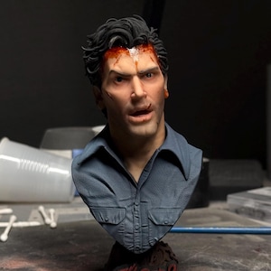 May include: A detailed resin bust of a man with dark hair, wearing a blue collared shirt. The figure features realistic skin tones, blood stains on the forehead, and a textured base with the red text Evil Dead 2 Dead by Dawn printed on the front.