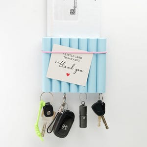 Könnte beinhalten: Ein hellblauer Schlüsselhalter mit einem rosa Gummiband, das eine Karte mit der Aufschrift "A little card to say a big thank you" hält. Vier Schlüsselsätze hängen von unten, darunter ein Autoschlüssel und ein Schlüssel mit einem neongrünen Schlüsselband.