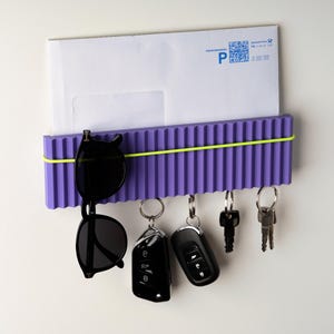 May include: A purple wall-mounted organizer with a ribbed design, holding a white envelope, sunglasses, and keys. A yellow elastic band secures the items. The organizer has key hooks and is designed for home or office use.