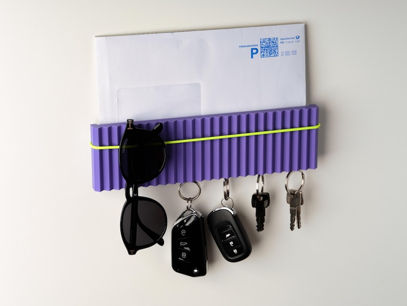 May include: A purple wall-mounted organizer with a ribbed design, holding a white envelope, sunglasses, and keys. A yellow elastic band secures the items. The organizer has key hooks and is designed for home or office use.