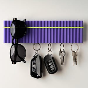 May include: A purple key holder with a neon yellow accent holds black sunglasses and several sets of keys. The key holder has a ribbed design and is mounted on a white wall.