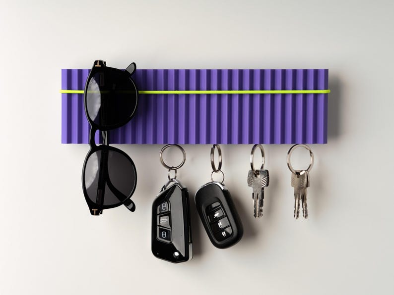 May include: A purple key holder with a neon yellow accent holds black sunglasses and several sets of keys. The key holder has a ribbed design and is mounted on a white wall.