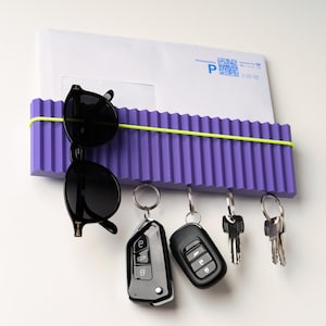 May include: A purple wall-mounted organizer with a corrugated design, holding a white envelope, black sunglasses, and keys. A neon green elastic band secures the items. The keys include car remotes and traditional keys.