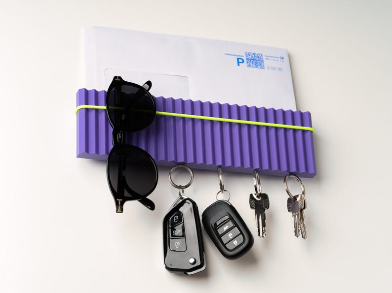 May include: A purple wall-mounted organizer with a corrugated design, holding a white envelope, black sunglasses, and keys. A neon green elastic band secures the items. The keys include car remotes and traditional keys.