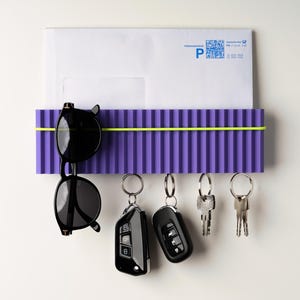 May include: A purple wall-mounted organizer with a yellow band, holding a white envelope, black sunglasses, and keys. The organizer has a ribbed design. The keys include car key fobs and traditional keys.