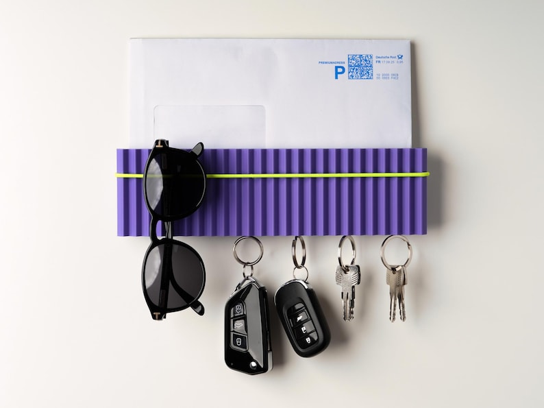 May include: A purple wall-mounted organizer with a yellow band, holding a white envelope, black sunglasses, and keys. The organizer has a ribbed design. The keys include car key fobs and traditional keys.