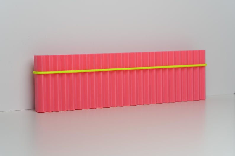 May include: A rectangular, pink container with vertical ridges and a neon yellow band wrapped around it. The container is set against a white background, creating a minimalist aesthetic. The object appears to be a storage box or organizer.