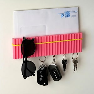 Op de afbeelding: Een roze wandorganizer met een gele elastiek, met een witte envelop, zwarte zonnebril en sleutels. De organizer heeft een geribbeld ontwerp en haken voor sleutels.