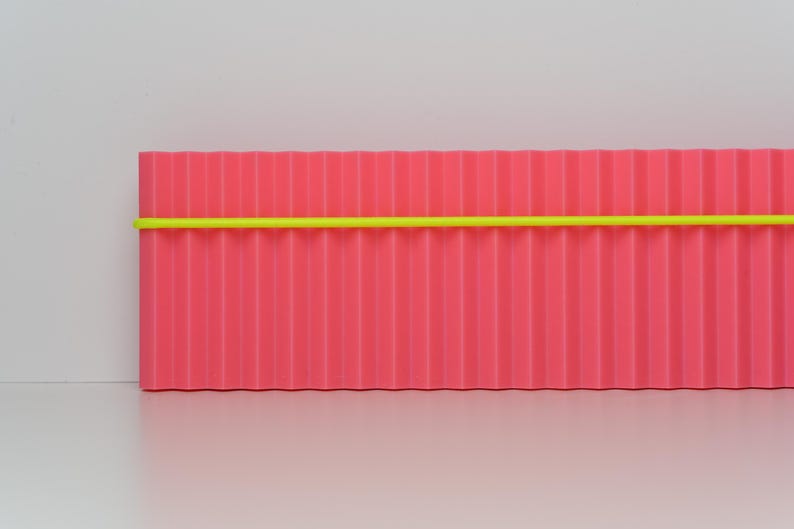 May include: A rectangular, corrugated, pink object with a bright yellow horizontal stripe. The object is set against a white background. The design is minimalist and modern.