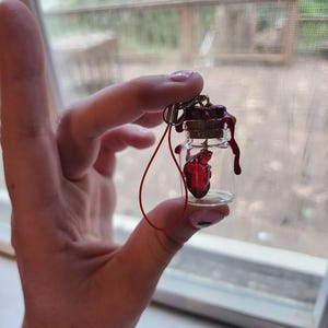 Peut inclure: Un petit flacon en verre avec un bouchon en liège et un objet rouge en forme de cœur à l'intérieur. Le bouchon est orné d'un effet de cire rouge foncé dégoulinante. Une ficelle rouge est attachée au flacon.