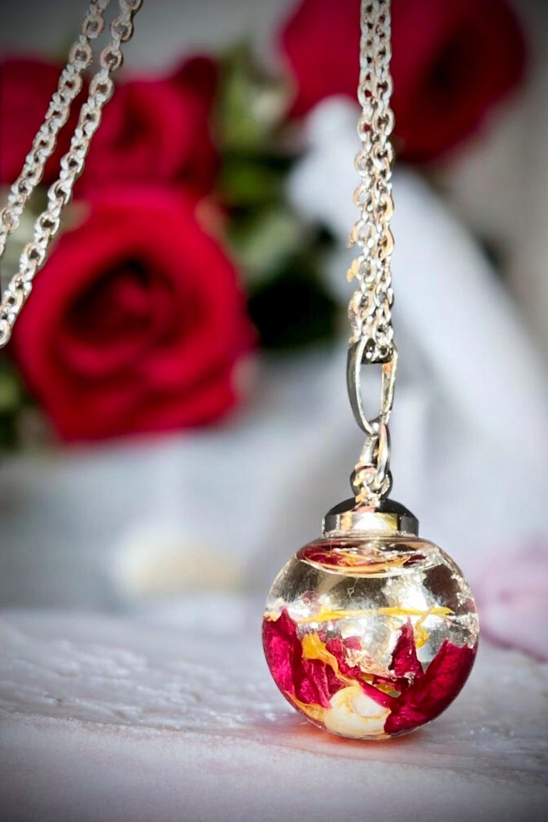 May include: Silver chain necklace with a clear, round pendant. The pendant is filled with red and yellow flower petals and gold flakes. The background features a blurred image of red roses.