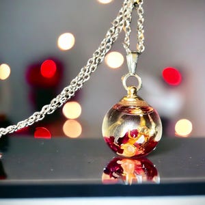 May include: A silver chain necklace with a clear, spherical pendant. The pendant contains red and gold floral elements. The background features blurred red and white lights, creating a bokeh effect. The pendant is reflected on a black surface.