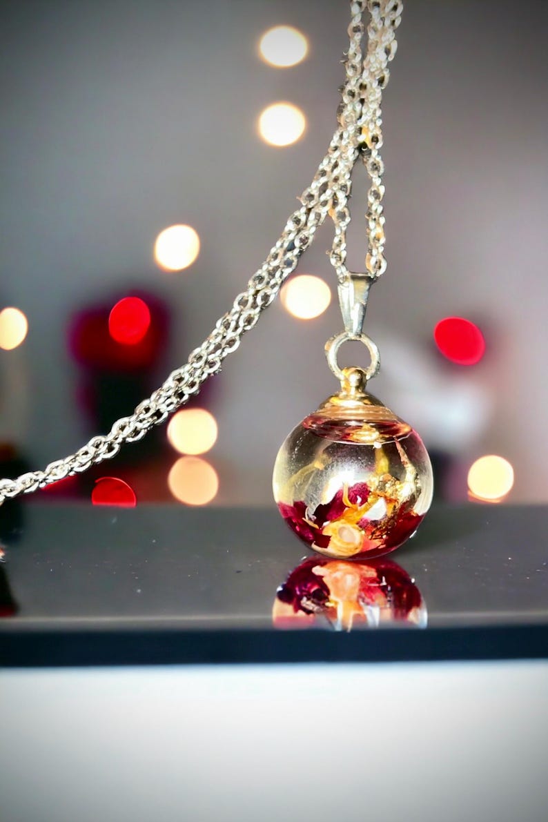 May include: A silver chain necklace with a clear, spherical pendant. The pendant contains red and gold floral elements. The background features blurred red and white lights, creating a bokeh effect. The pendant is reflected on a black surface.