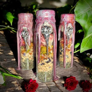 May include: Three glass bottles filled with dried herbs and flowers, sealed with pink wax and topped with a silver bird skull. The bottles are arranged on a weathered wooden surface, with red roses in the foreground. The bottles are filled with a variety of dried herbs and flowers.