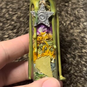 May include: A small glass bottle filled with dried herbs and flowers, sealed with green wax and a silver star charm. The bottle contains yellow, purple, and green botanicals.