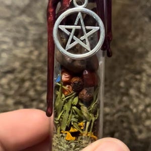 May include: A small glass vial filled with various dried herbs, seeds, and other natural elements. The vial is sealed with a dark red wax and topped with a silver pentagram charm. The contents are visible through the clear glass.