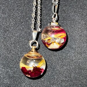 May include: Two silver chain necklaces each feature a clear, spherical pendant. The pendants contain red and yellow floral elements, with silver and gold accents. The necklaces are displayed against a dark background.