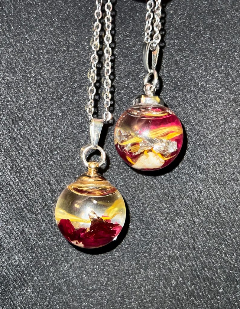 May include: Two silver chain necklaces each feature a clear, spherical pendant. The pendants contain red and yellow floral elements, with silver and gold accents. The necklaces are displayed against a dark background.