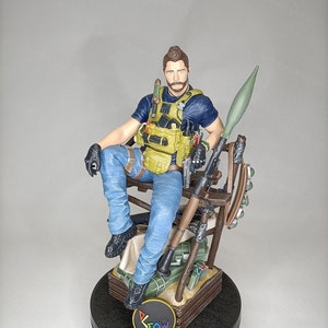 Könnte beinhalten: Eine detaillierte Figur eines Soldaten, der auf einer Holzkiste mit verschiedenen militärischen Ausrüstungsgegenständen und Waffen sitzt. Der Soldat trägt ein blaues Hemd, eine gelbe Weste und eine blaue Jeans. Die Figur steht auf einem schwarzen Sockel mit dem Wort "Drow" in Regenbogenfarben.