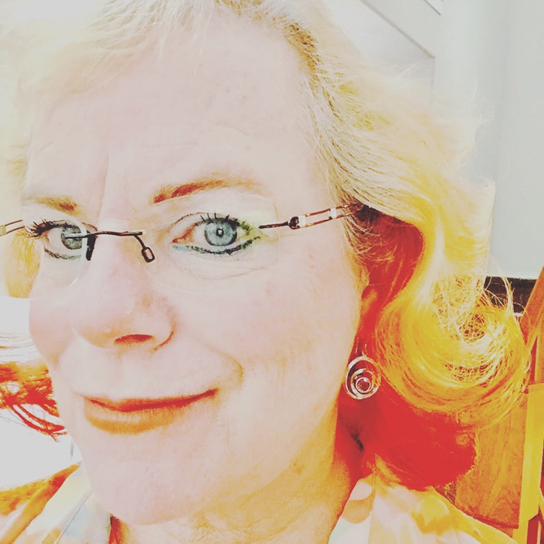 May include: A close-up portrait of a person with blonde hair wearing glasses and a small circular earring. The person is smiling and looking directly at the camera.