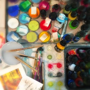 May include: A close-up of an artist's workspace with various art supplies, including paint, brushes, and a palette. The image shows a variety of colors, including red, blue, green, and yellow. There are also several containers of paint, some of which are open and some of which are closed. The image is a great example of the tools and materials that artists use to create their work.