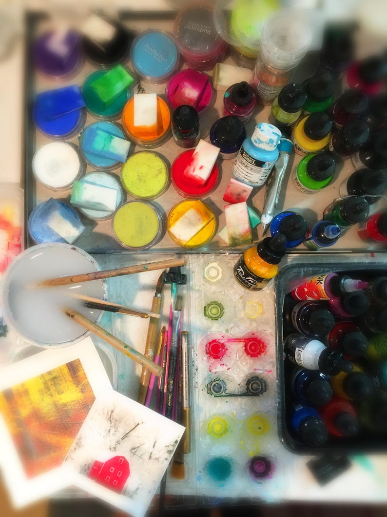 May include: A close-up of an artist's workspace with various art supplies, including paint, brushes, and a palette. The image shows a variety of colors, including red, blue, green, and yellow. There are also several containers of paint, some of which are open and some of which are closed. The image is a great example of the tools and materials that artists use to create their work.
