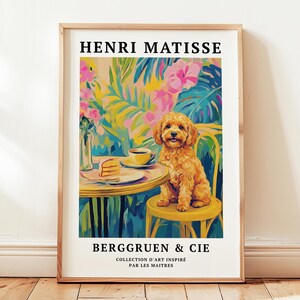 May include: A framed art print depicting a golden dog at a table with coffee and cake, set against a backdrop of vibrant tropical plants. The artwork includes the text "HENRI MATISSE" and "BERGGRUEN & CIE". The print is in a wooden frame.