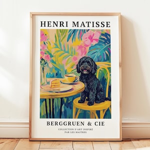May include: A framed art print featuring a black dog sitting on a yellow chair at a table with a cup of coffee and cake. The background is a colorful tropical scene. The print includes the text "HENRI MATISSE" and "BERGGRUEN & CIE".